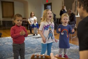 A warm, energetic early childhood music class where young children play drums, shakers, and scarves together. The teacher leads them in a lively activity, and the children are fully engaged, making eye contact, moving their bodies, and interacting with real instruments instead of screens.