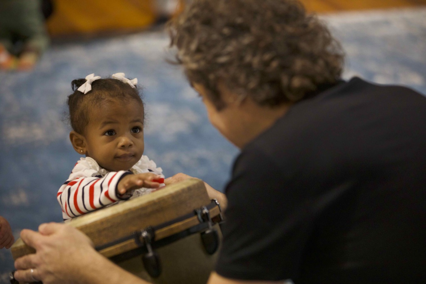 A baby reaching for a small drum while a grown-up holds it close