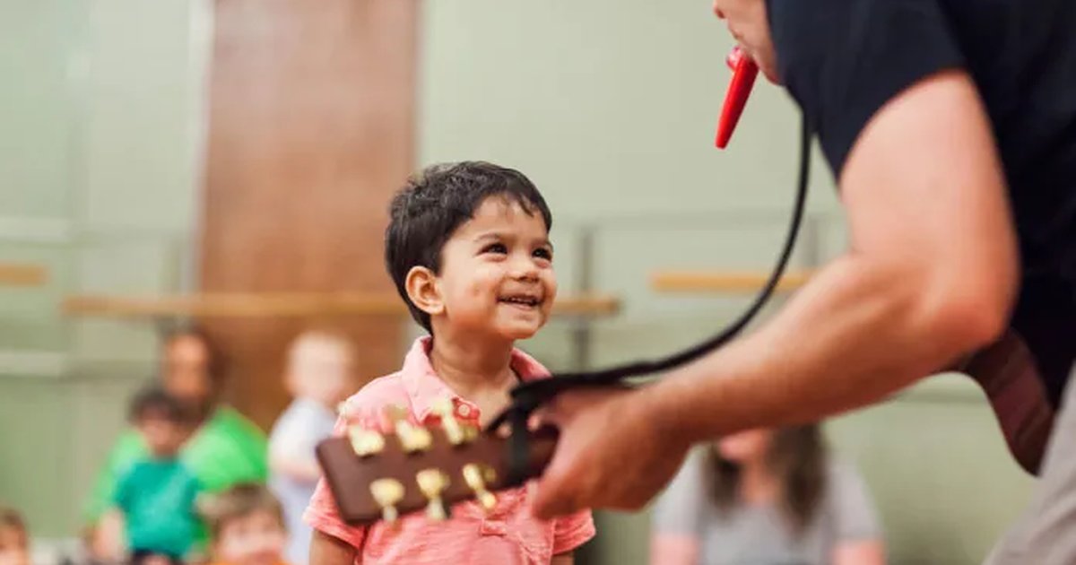 A boy grinning at a guitar while a teacher plays the kazoo