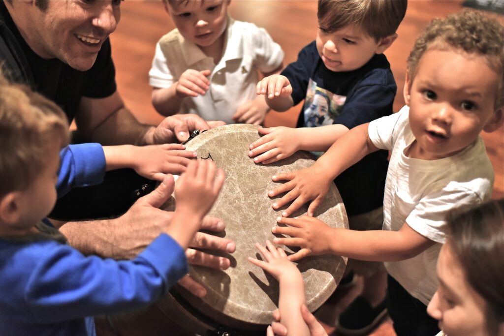 Toddlers gathered around the drum banging on it with the preschool music teacher.